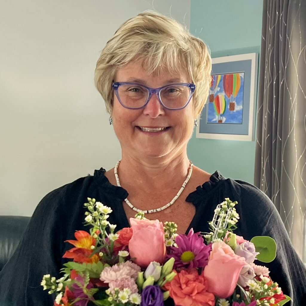 Dr. Melissa Sadin smiling with short blonde hair and purple glasses, holding a vibrant bouquet of pink roses and purple flowers.
